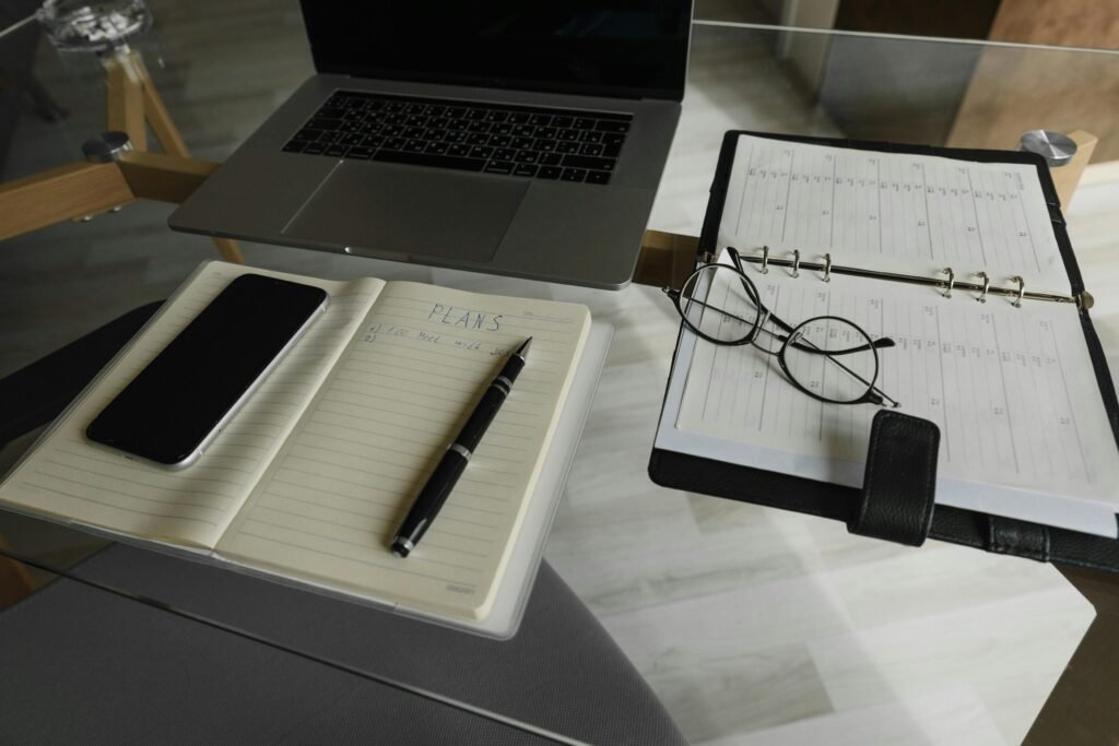pexels-photo-5710681-5710681 A neatly organized workspace featuring a laptop, smartphone, notebook, pen, and planner on a glass table.